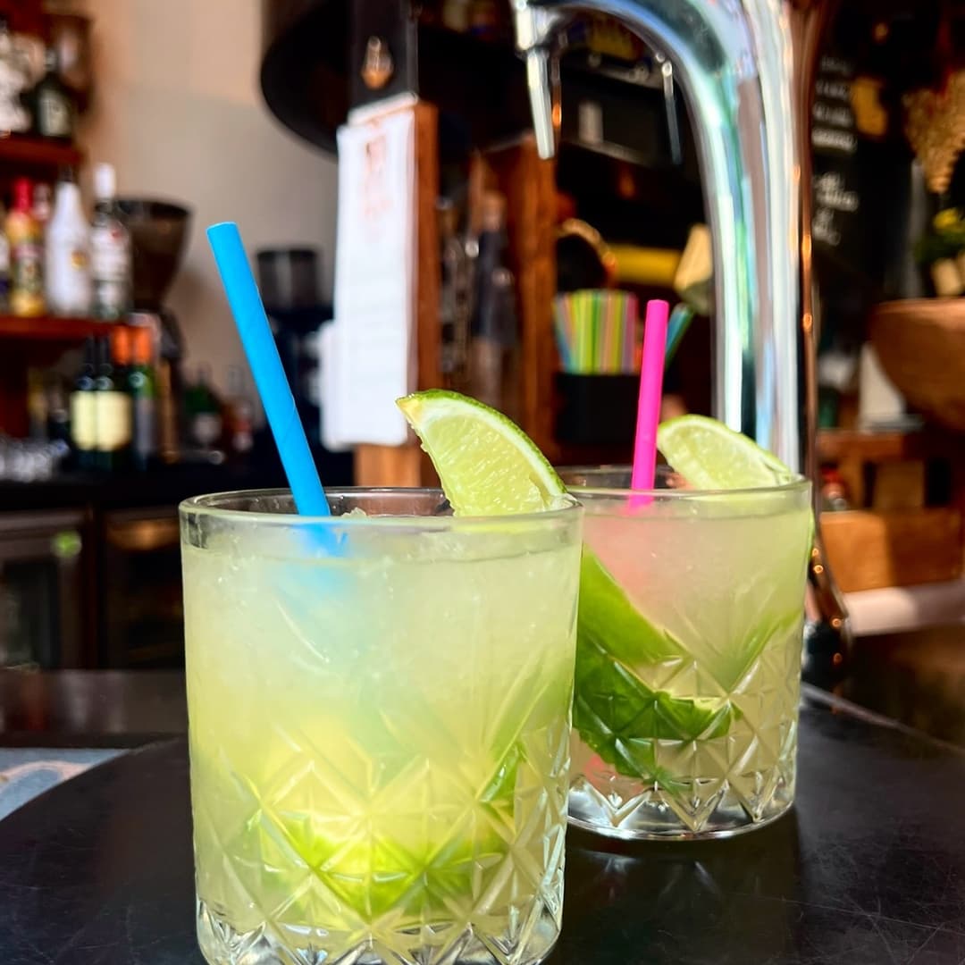 Two refreshing cocktails with lime wedges and colorful straws, set against a vibrant bar backdrop.