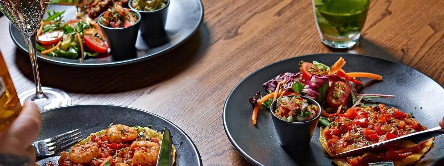 Plates of vibrant dishes featuring shrimp, fresh salads, and colorful garnishes on a wooden table, with drinks nearby.