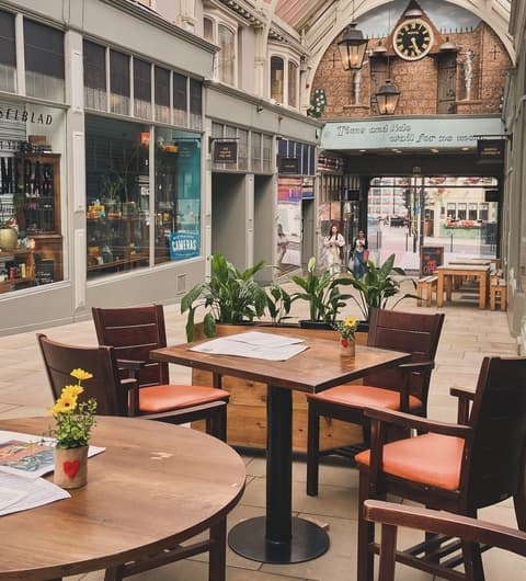 Charming outdoor dining area with wooden tables, potted plants, and a clock above in a vibrant, covered arcade.