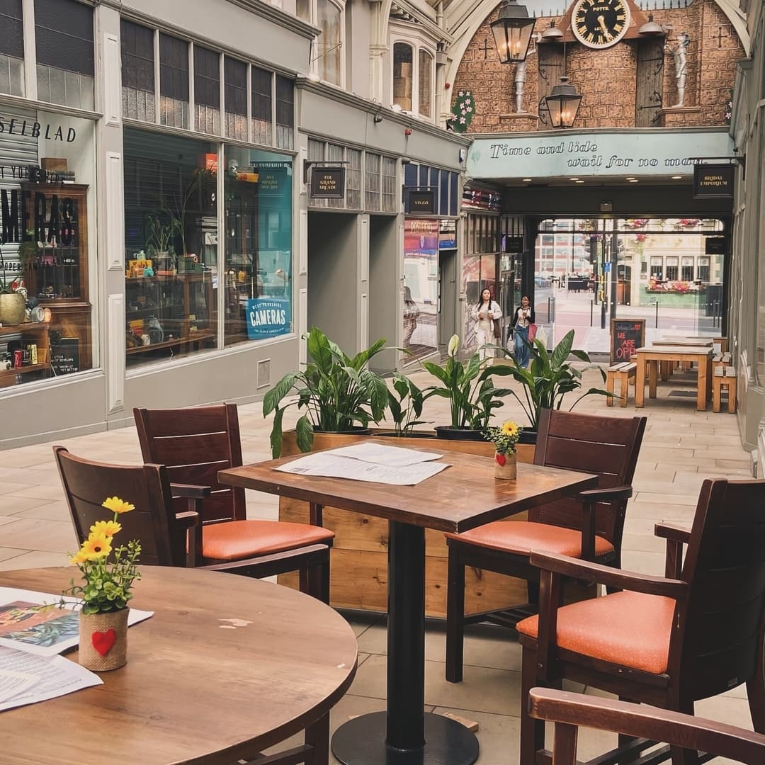 Charming outdoor dining area with wooden tables, orange cushioned chairs, and potted plants under a historic archway.