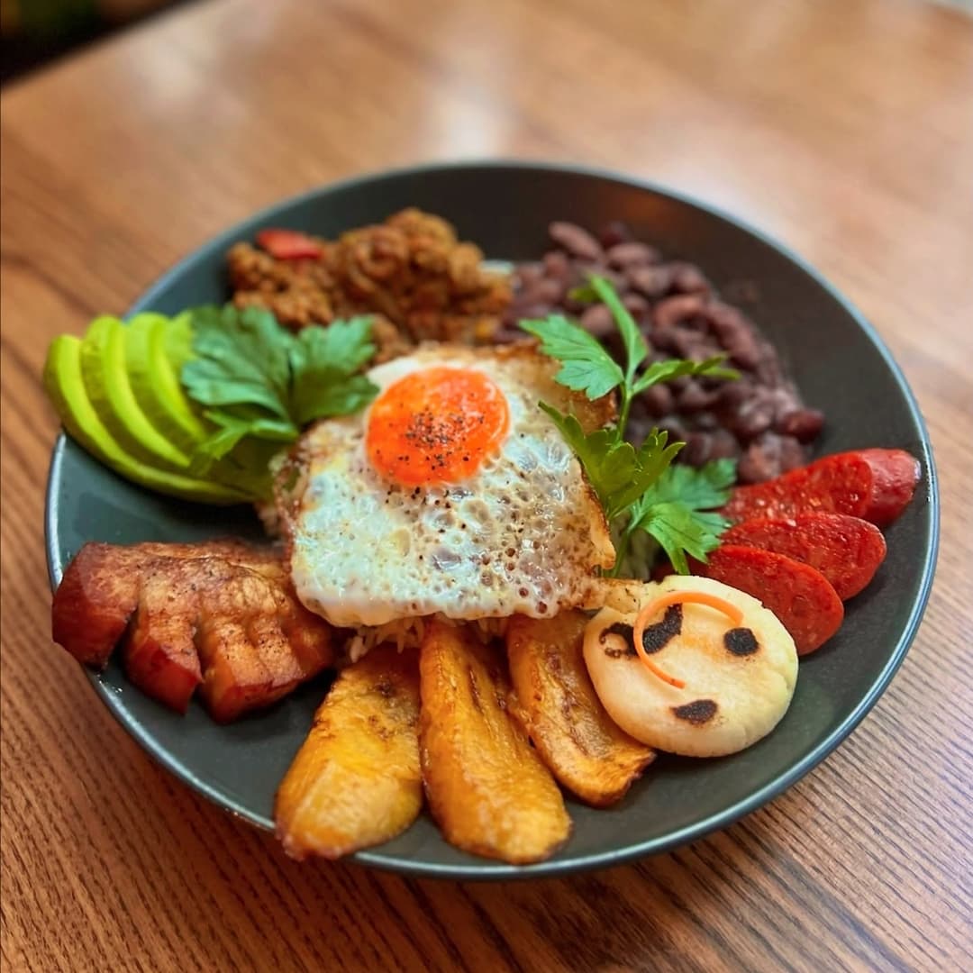 Colorful plate featuring fried egg, plantains, avocado, beans, chorizo, and a smiling face made of dough.