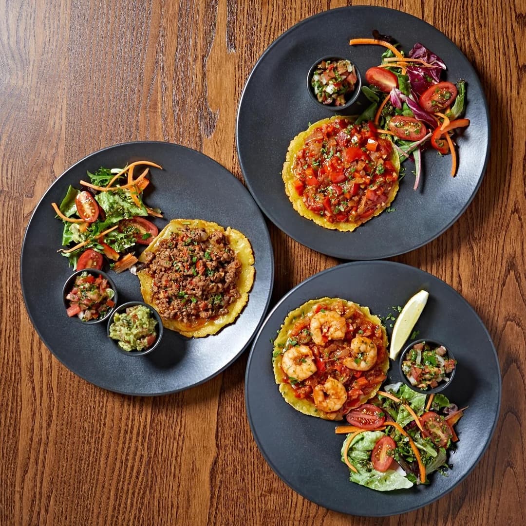 Three vibrant plates of colorful tacos and salads, featuring fresh ingredients and garnishes, on a wooden table.