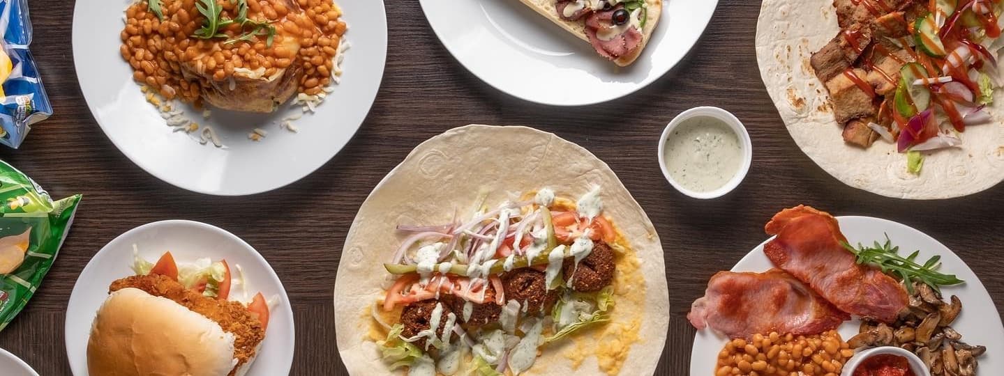 A spread of diverse dishes including tacos, sandwiches, baked beans, and crispy bacon on white plates, set on a wooden table.