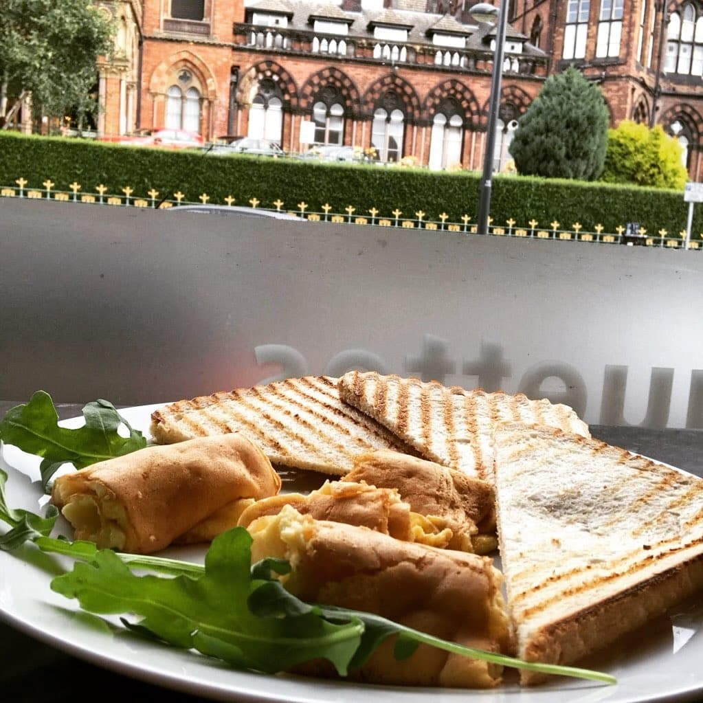 Grilled sandwiches and spring rolls on a plate, with fresh arugula, framed by a view of historic architecture outside.