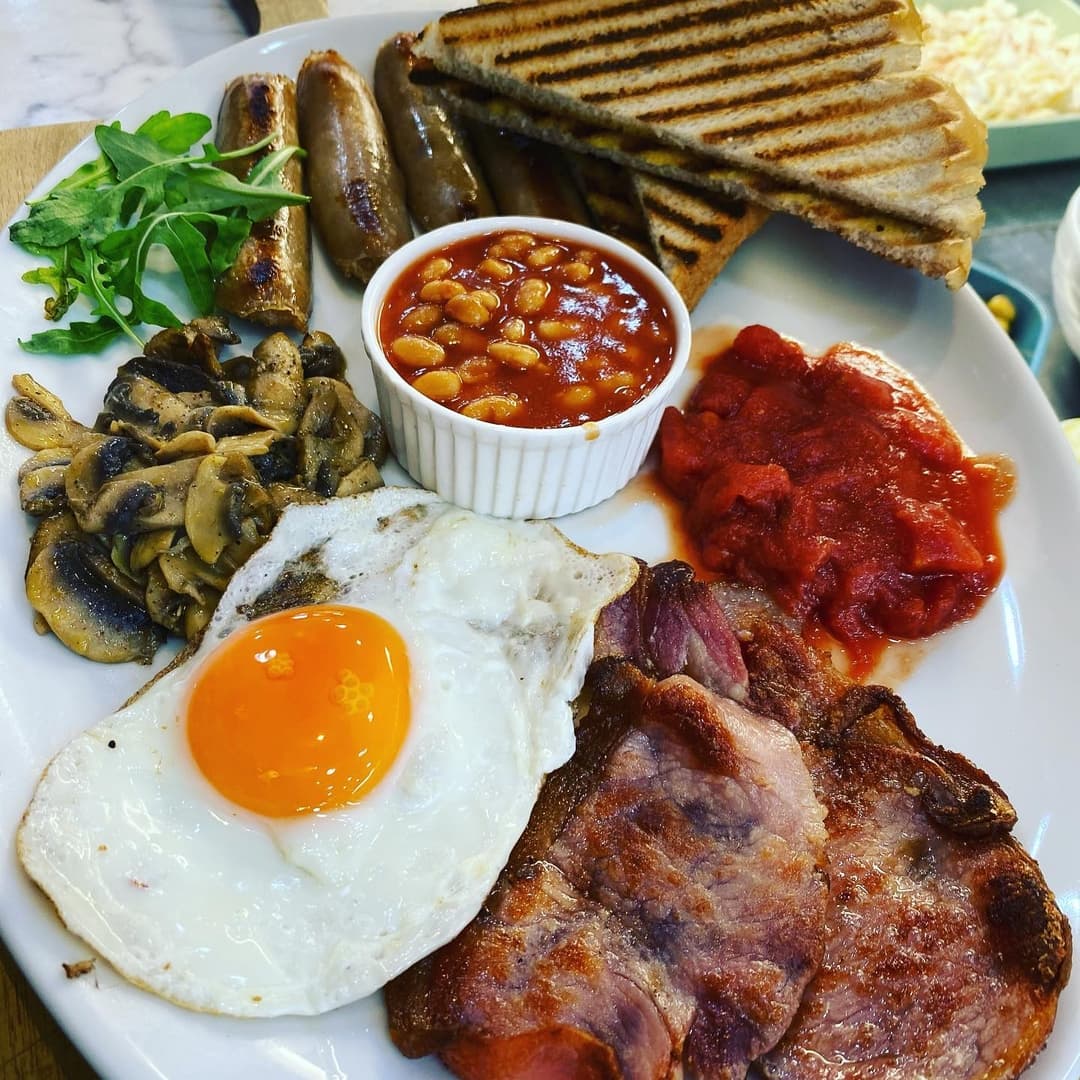 A hearty breakfast plate featuring sausages, bacon, fried egg, baked beans, grilled mushrooms, and toast.