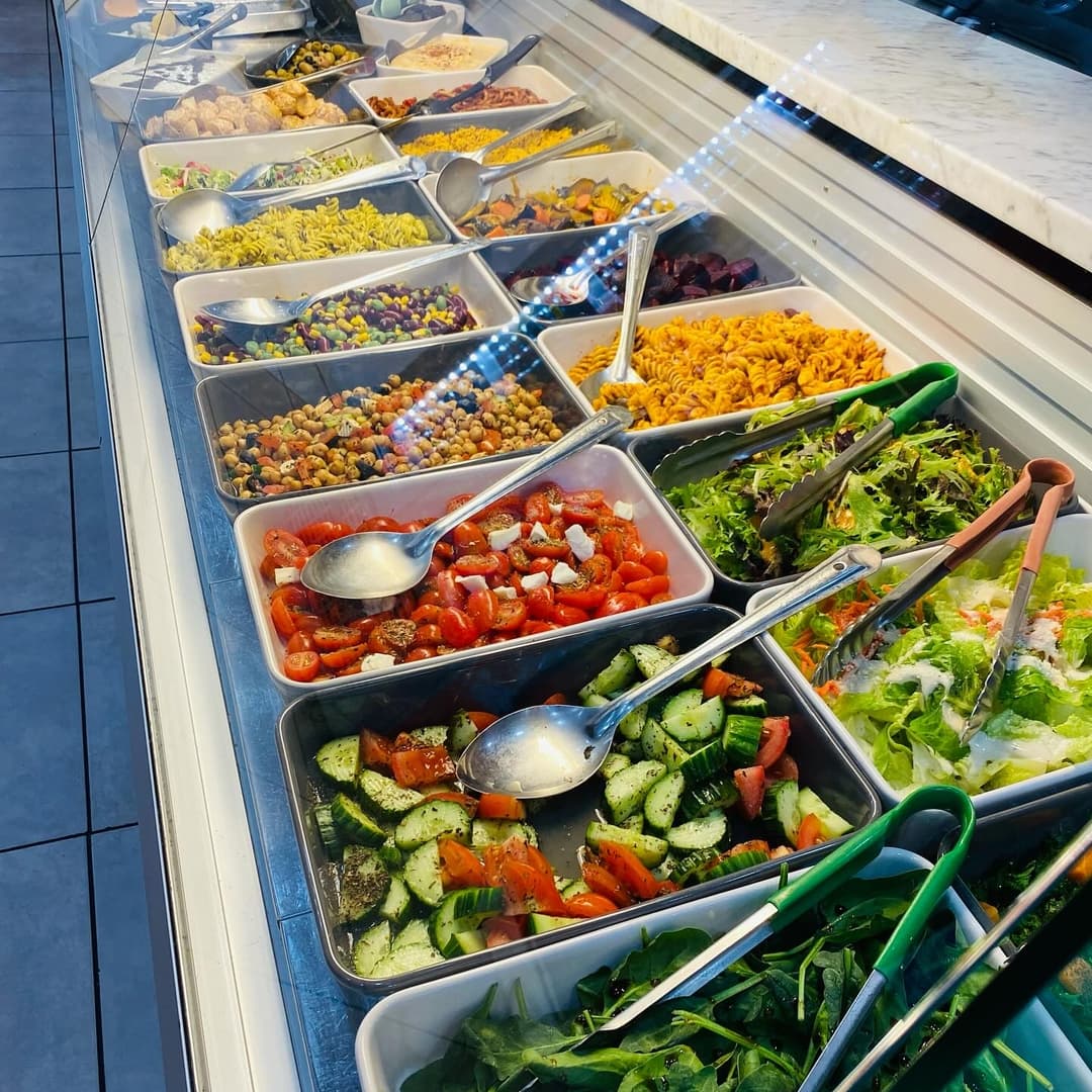 Colorful assortment of fresh salads and toppings in trays, with serving utensils, set in a bright, inviting deli display.
