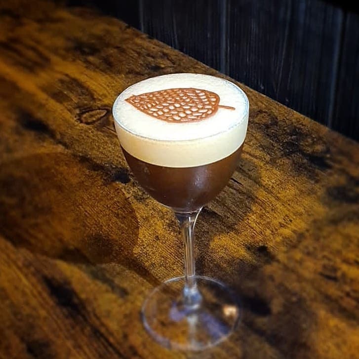A frothy cocktail with a leaf design on top, served in a elegant glass on a rustic wooden table.