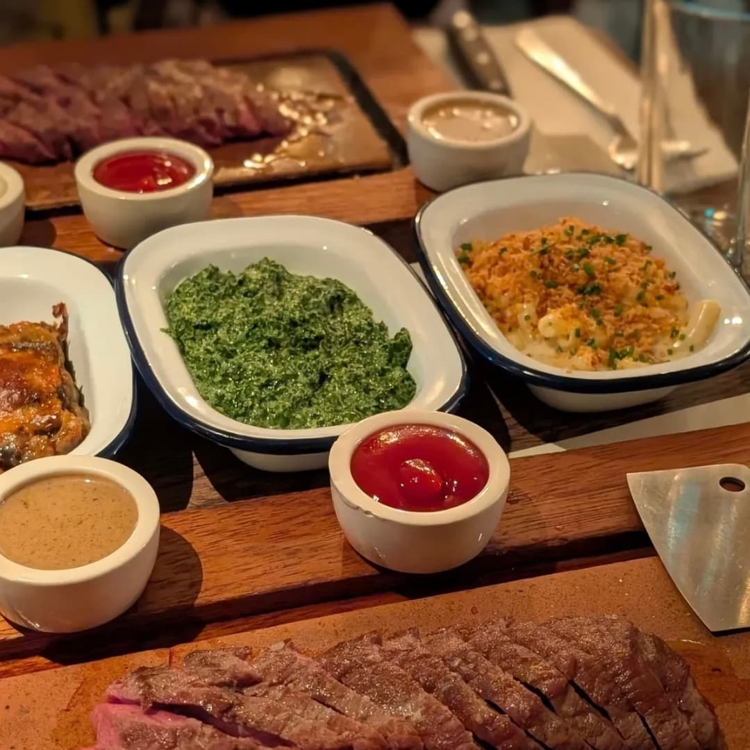 Plates of succulent steak, vibrant green spinach, creamy mac and cheese, and dipping sauces on a rustic wooden table.