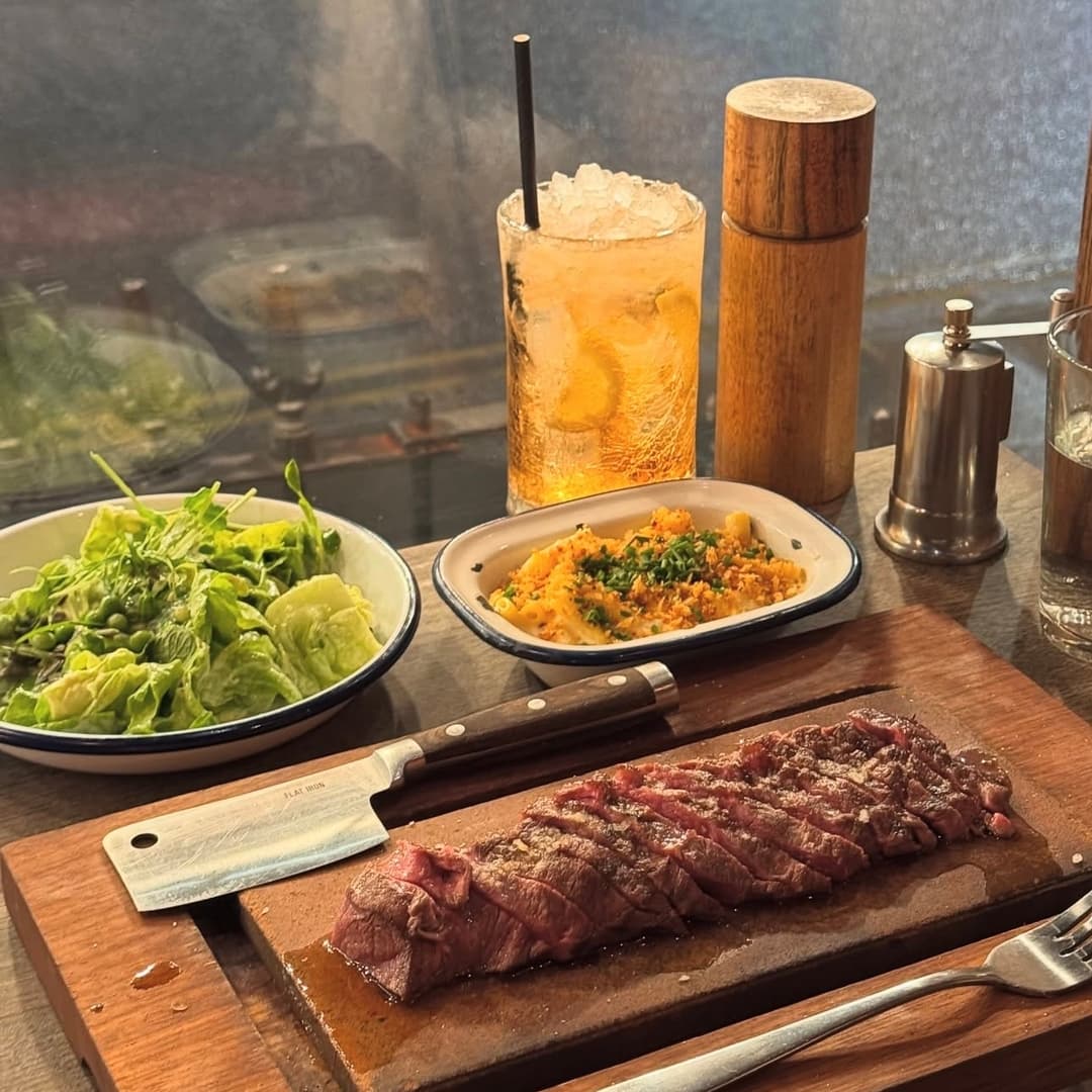 Juicy sliced steak on a wooden board, accompanied by a fresh salad, spiced rice, and a refreshing drink.