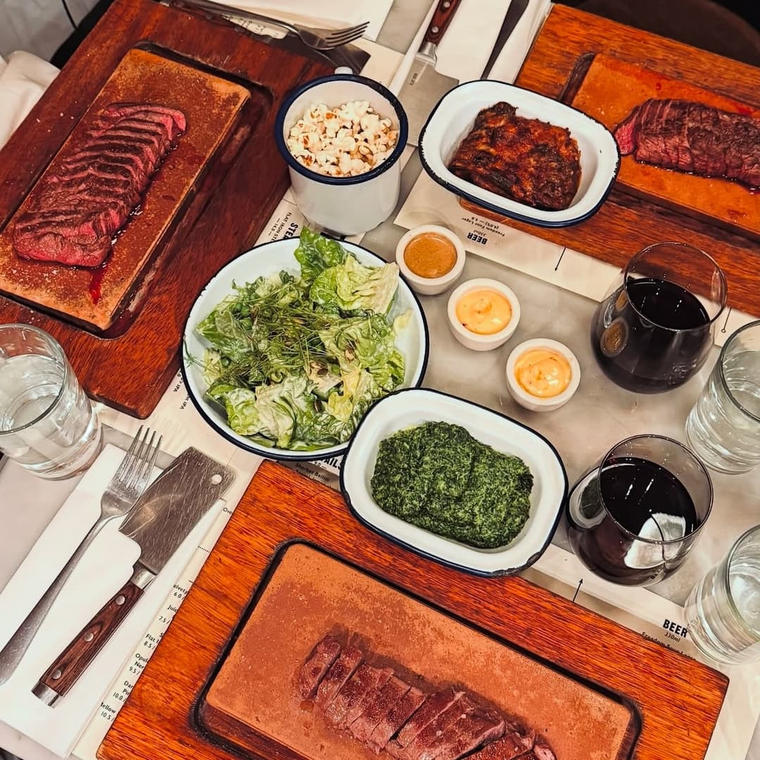 A vibrant dining table with various cuts of steak, fresh salad, sauces, and drinks, creating a lively atmosphere.