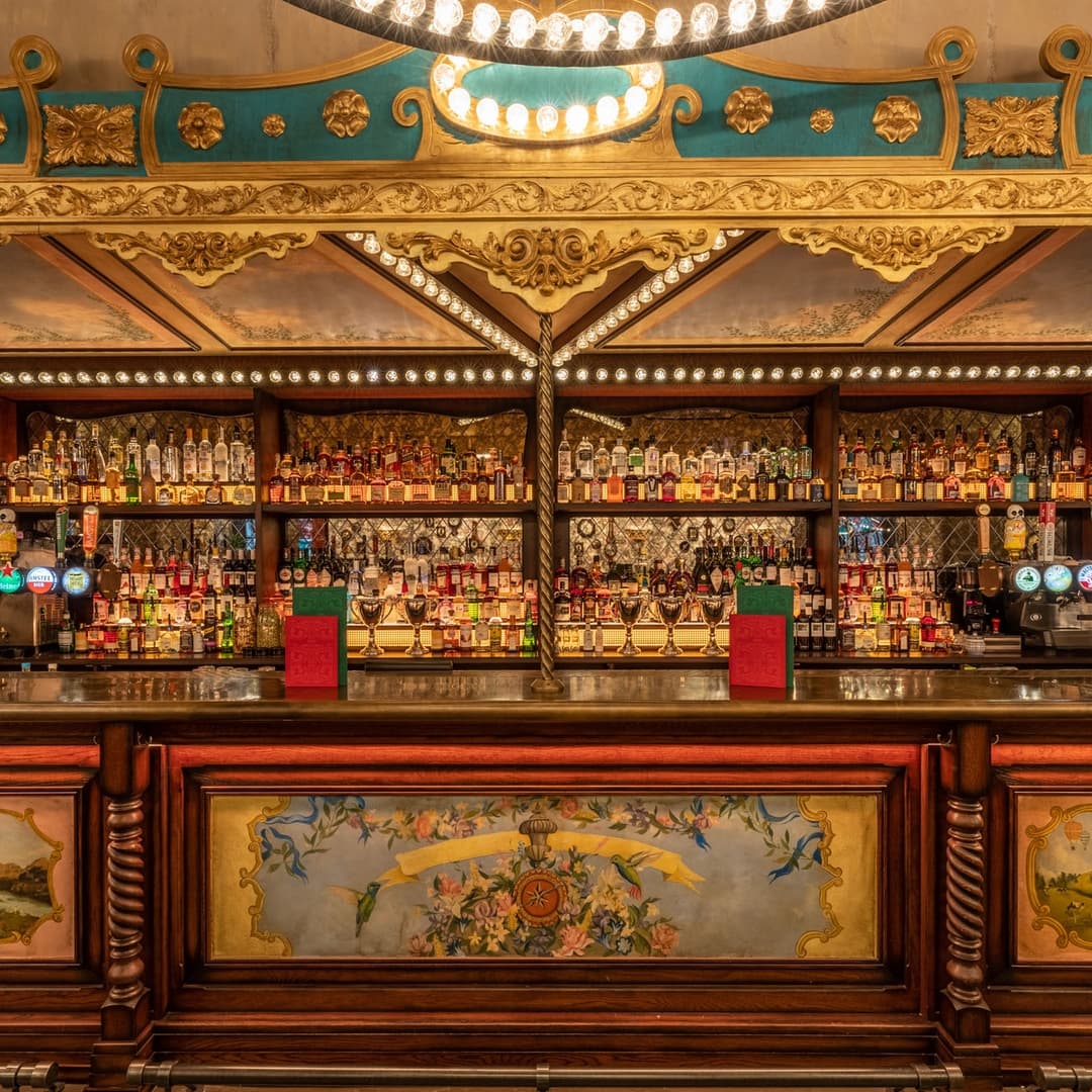 Elegant bar with ornate decor, shelves lined with colorful spirits, and warm lighting creating a vibrant atmosphere.