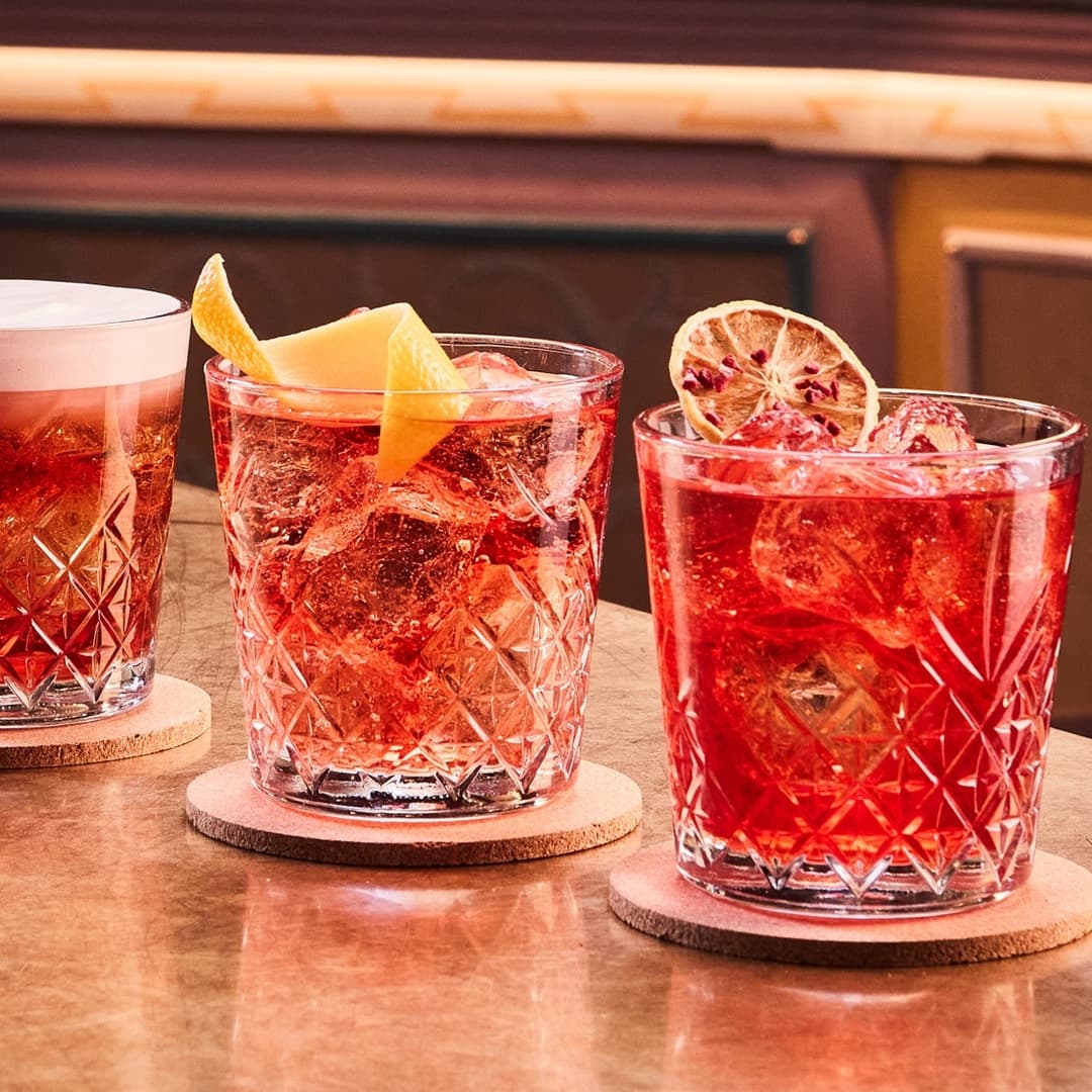 Three vibrant cocktails with citrus garnishes sit on a polished wooden table, showcasing a lively bar atmosphere.