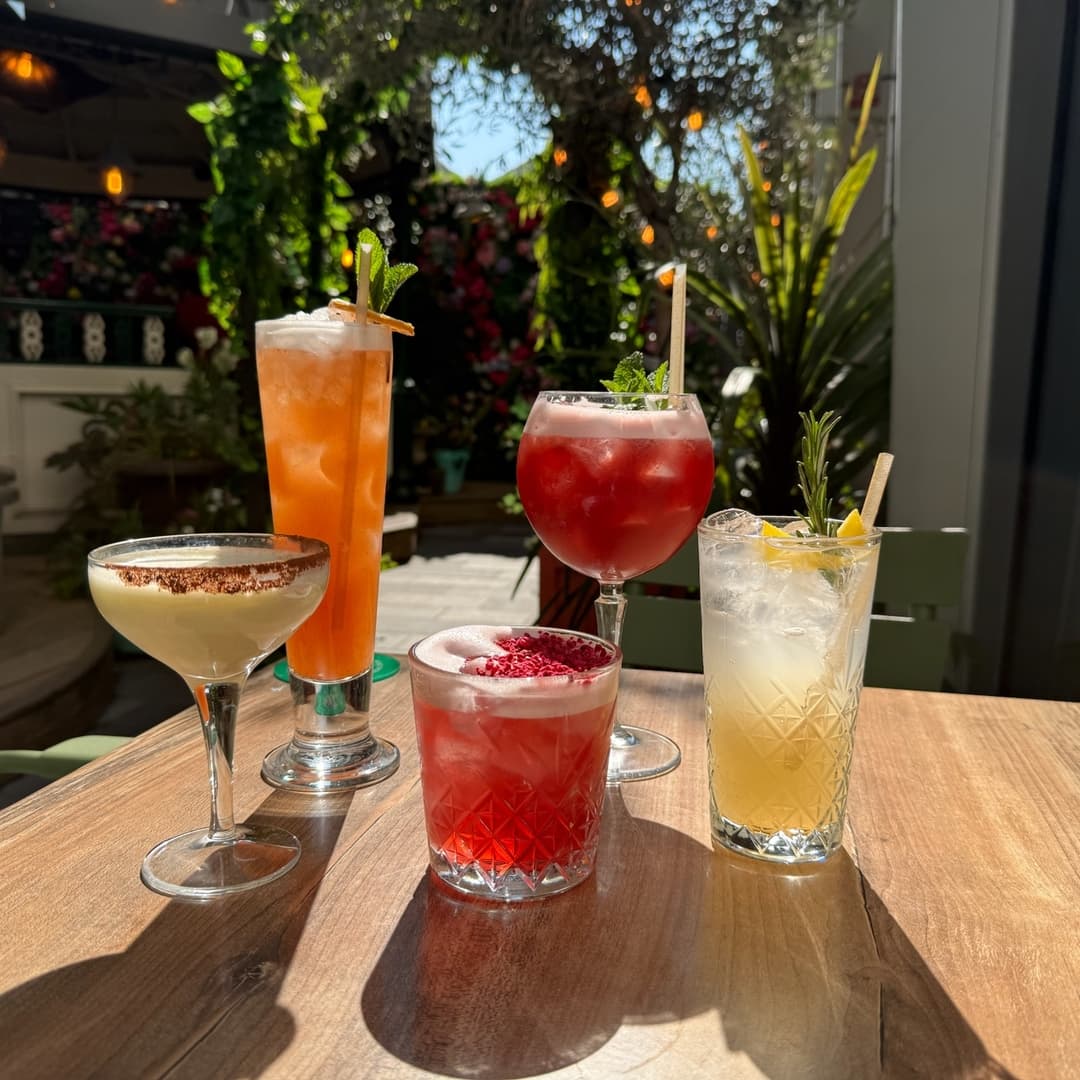 Five colorful cocktails on a wooden table, surrounded by lush greenery and warm sunlight, creating a vibrant atmosphere.
