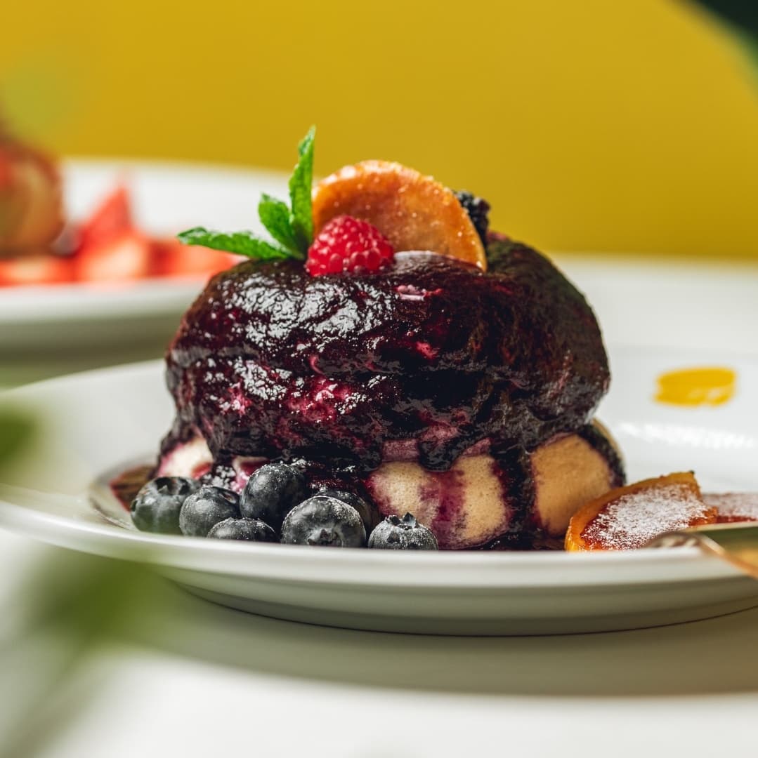 Fluffy pancake topped with dark berry compote, fresh blueberries, a raspberry, and a slice of orange, on a white plate.