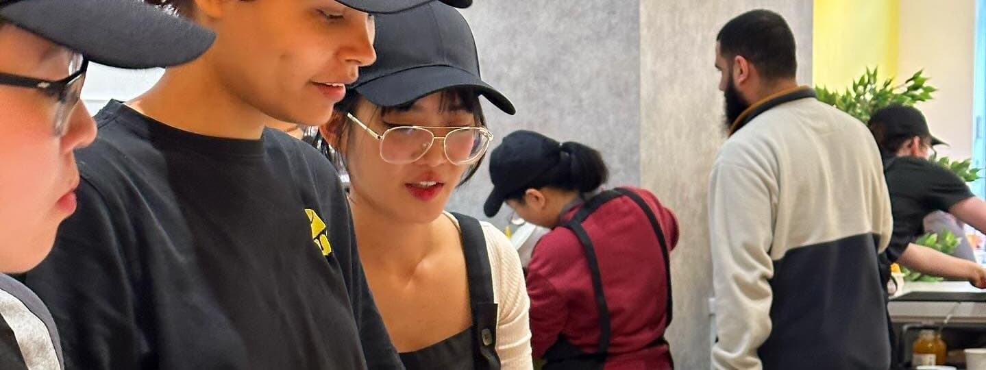 Staff members in black caps and aprons collaborate in a bright, modern kitchen, preparing food with focused expressions.