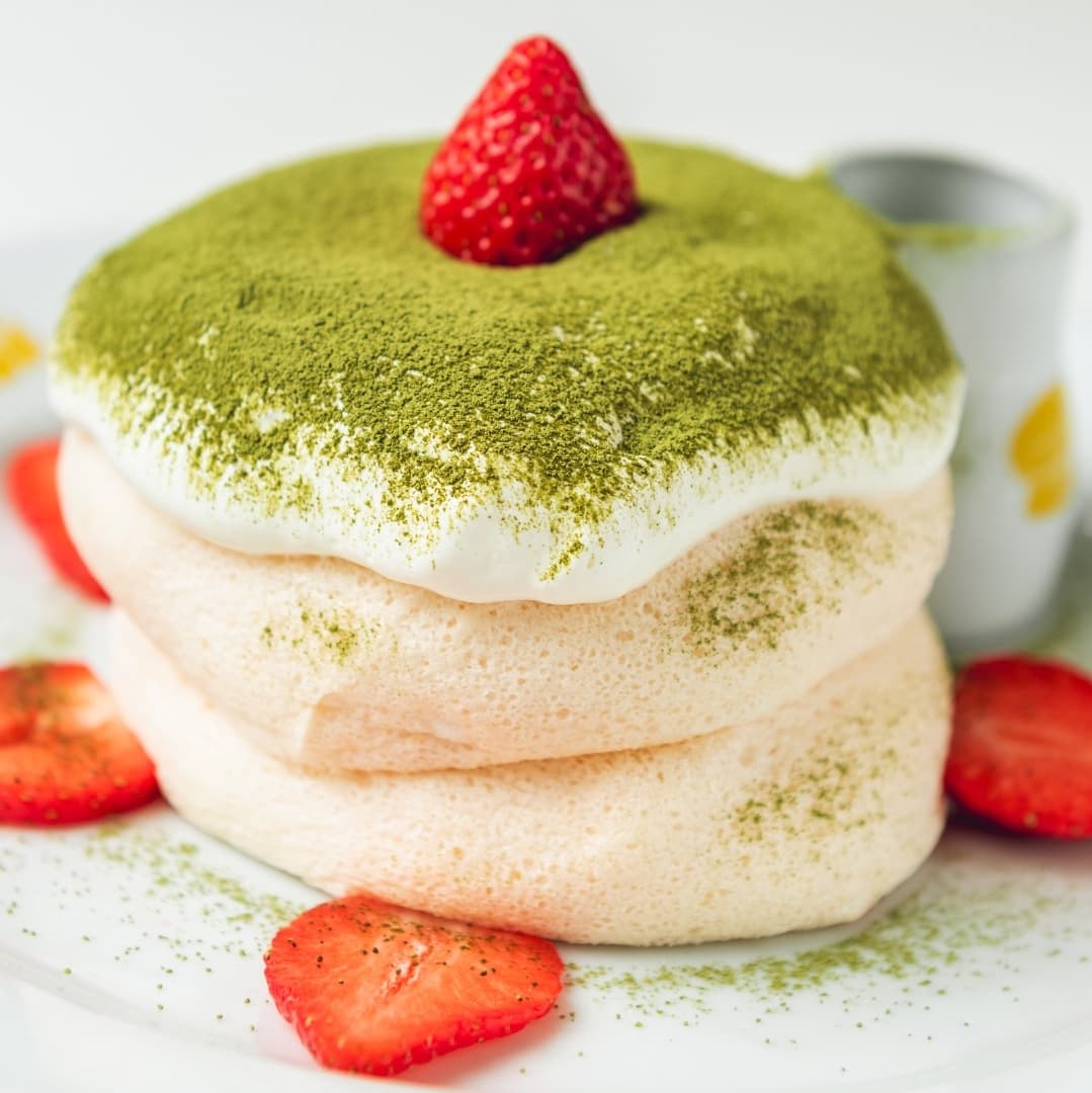Fluffy pancakes topped with matcha cream, a strawberry, and surrounded by fresh strawberry slices on a white plate.