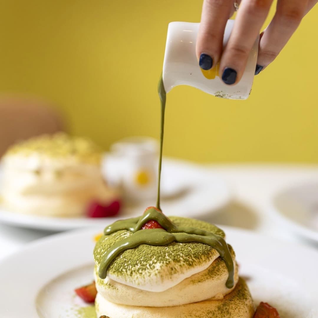 A hand drizzles green matcha sauce over fluffy pancakes, garnished with strawberries, against a bright yellow background.