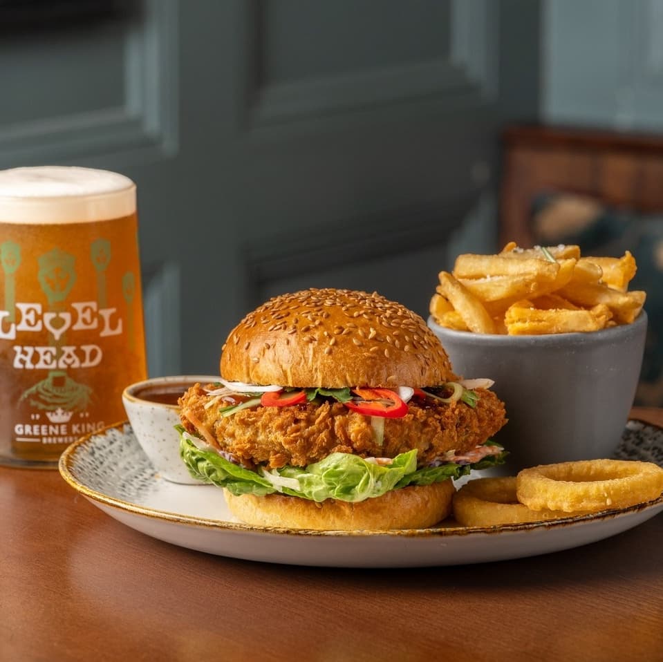 Crispy fried chicken burger with lettuce and chili, served with onion rings, fries, and a pint of beer on a wooden table.