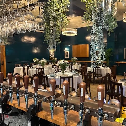 Elegant dining area with white tablecloths, floral centerpieces, and hanging greenery, complemented by a polished bar with beer taps.
