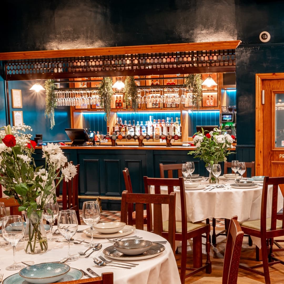 Elegant dining setup with white tablecloths, glassware, and a stylish bar adorned with greenery and bottles in the background.