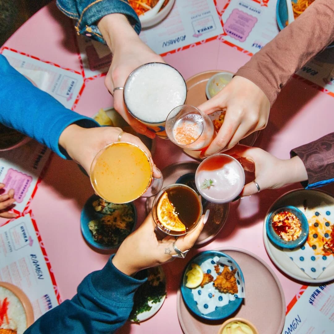 Colorful cocktails and drinks clink together over a pink table, surrounded by vibrant dishes and menus.