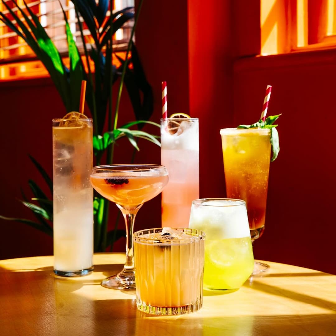 Colorful cocktails in various glasses arranged on a sunlit table, with vibrant orange walls and greenery in the background.