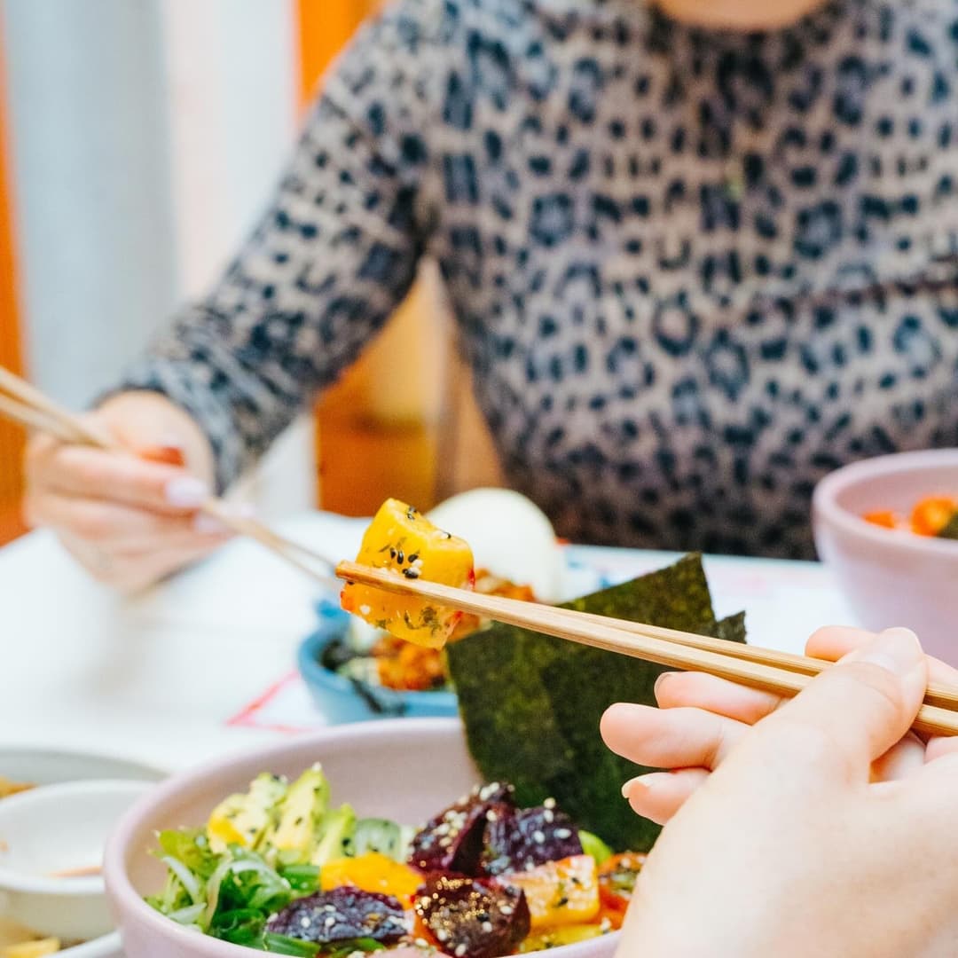 A hand holds chopsticks above a colorful bowl of sushi and fresh vegetables, with a cozy dining atmosphere.