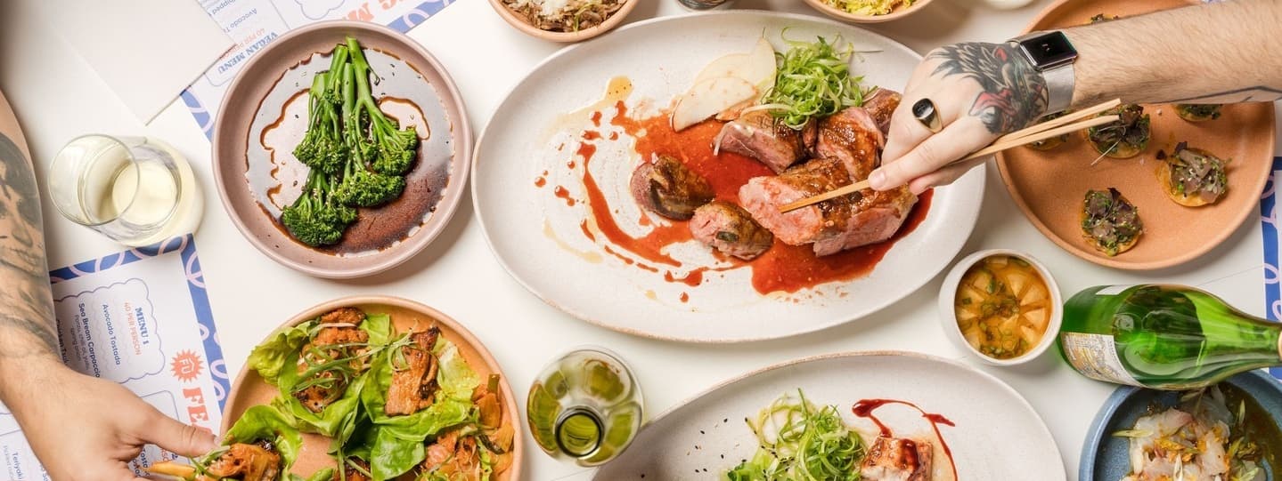 A vibrant spread of Asian dishes, including meats, greens, and sauces, with hands using chopsticks and drinks nearby.