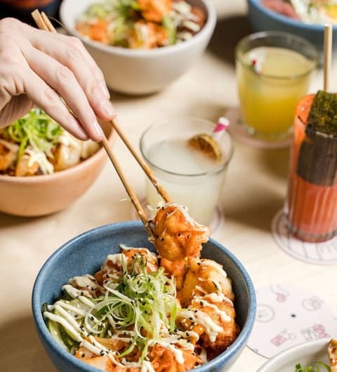 A hand using chopsticks to lift crispy tempura from a blue bowl, surrounded by colorful dishes and drinks.
