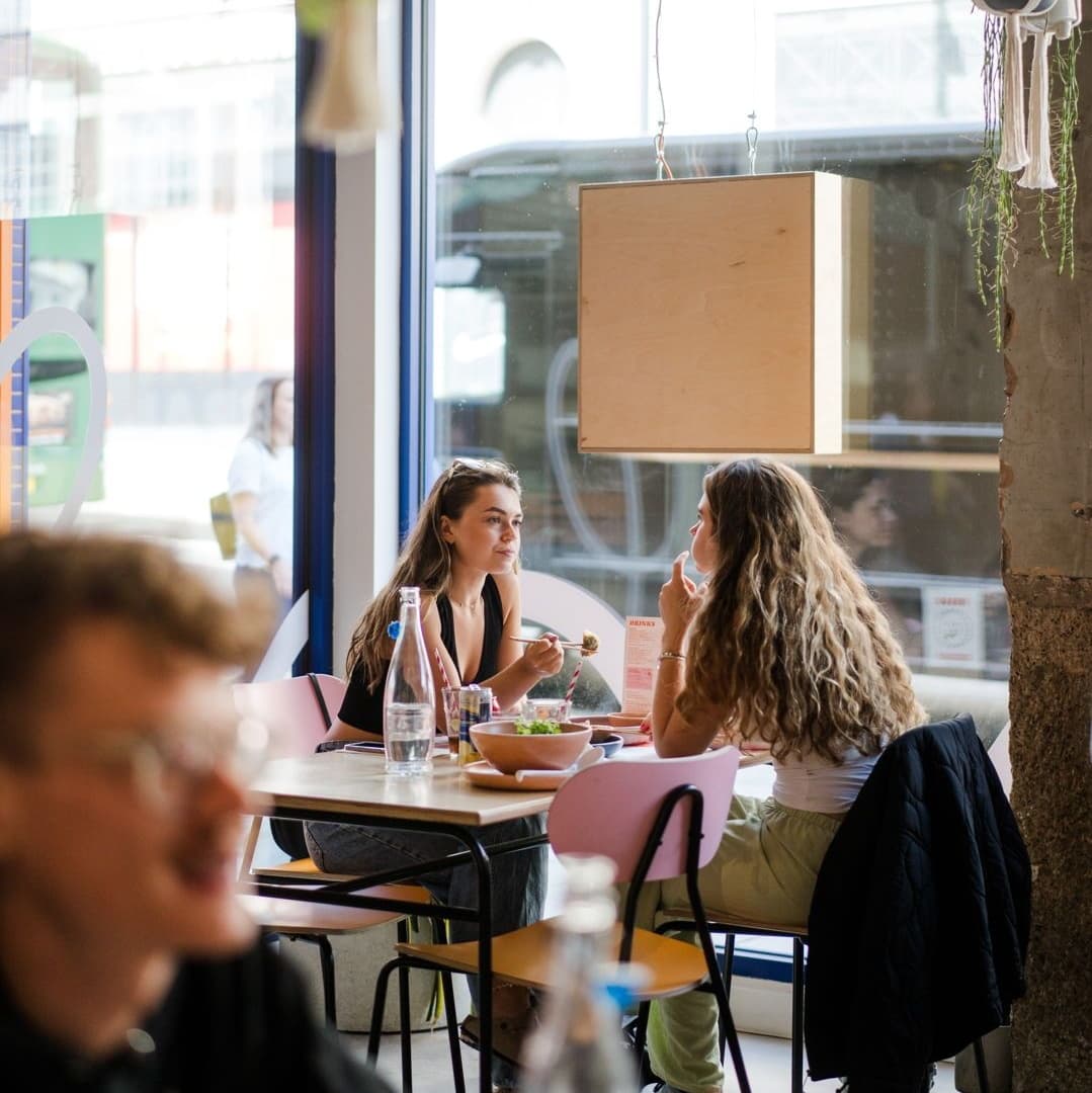 Two women share a meal at a modern table, surrounded by colorful decor and large windows letting in natural light.