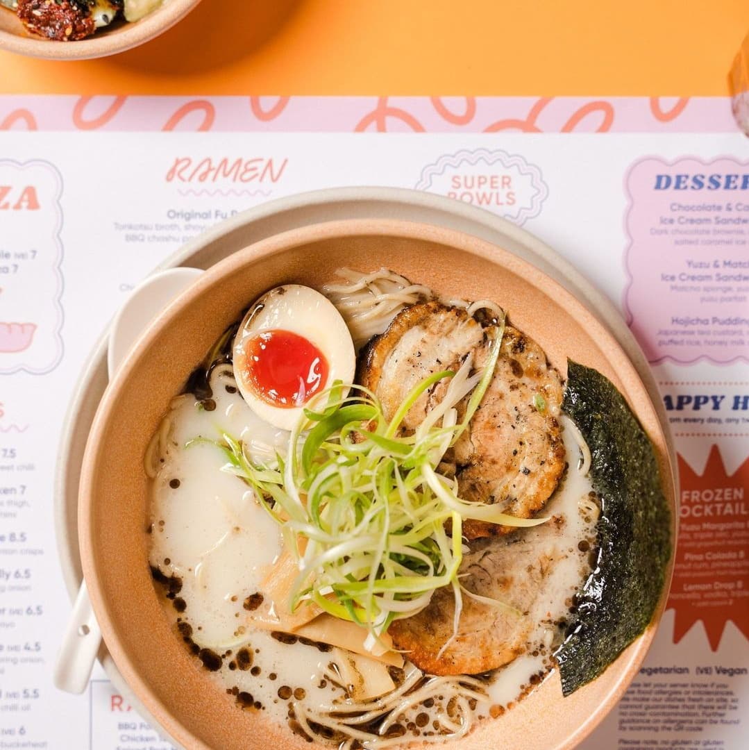 Bowl of ramen with sliced pork, soft-boiled egg, green onions, and seaweed, set on a vibrant orange table.