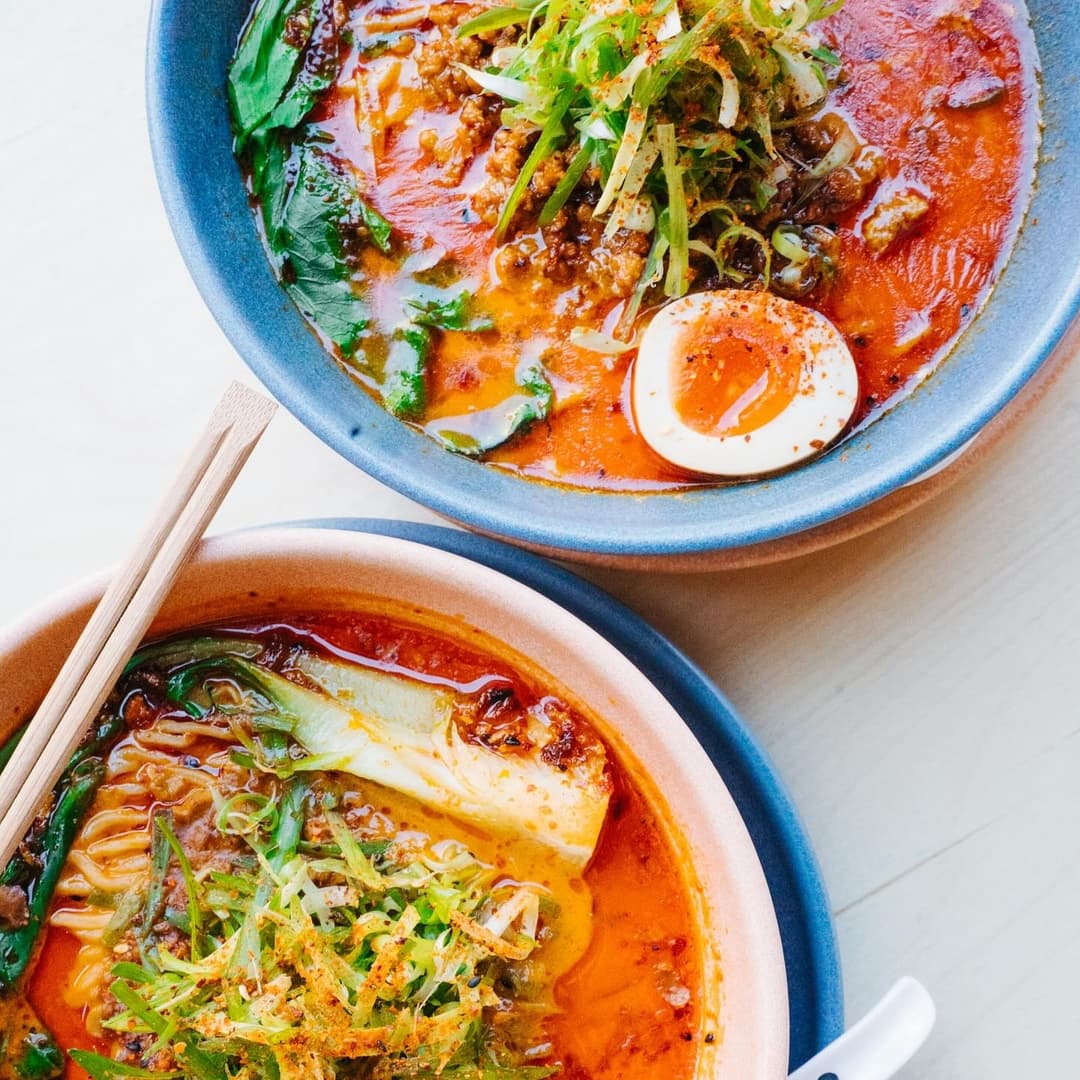 Two bowls of vibrant ramen, one topped with green onions and a soft-boiled egg, the other with leafy greens and spices.