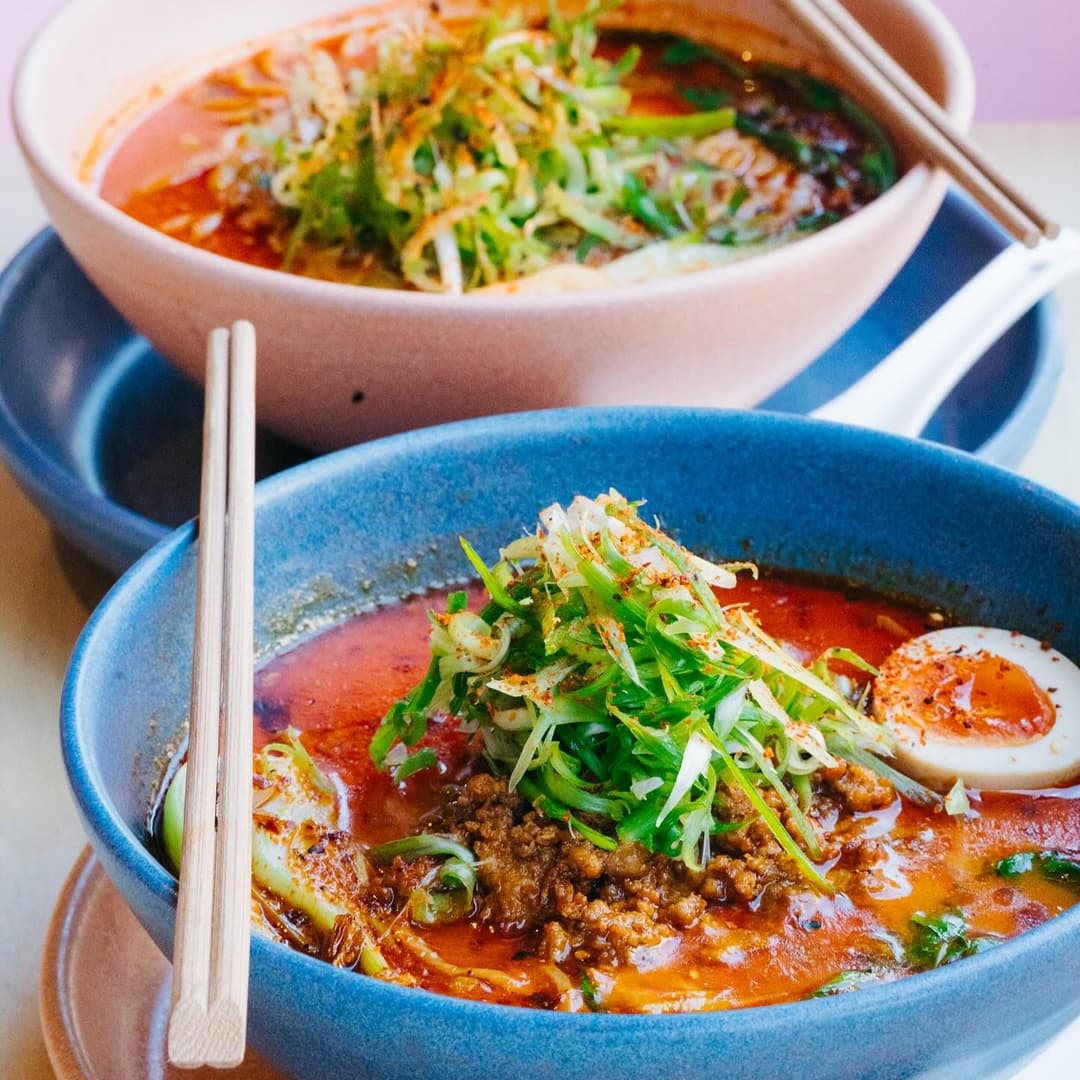 Two vibrant bowls of spicy ramen topped with green onions, minced meat, and a soft-boiled egg, set against a colorful backdrop.