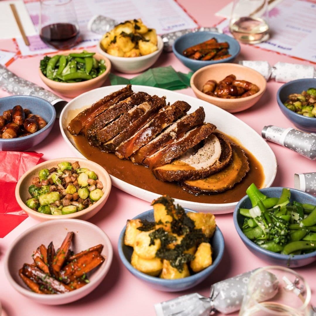 Sliced roast duck drizzled with sauce, surrounded by colorful bowls of vegetables and sides on a pink table.
