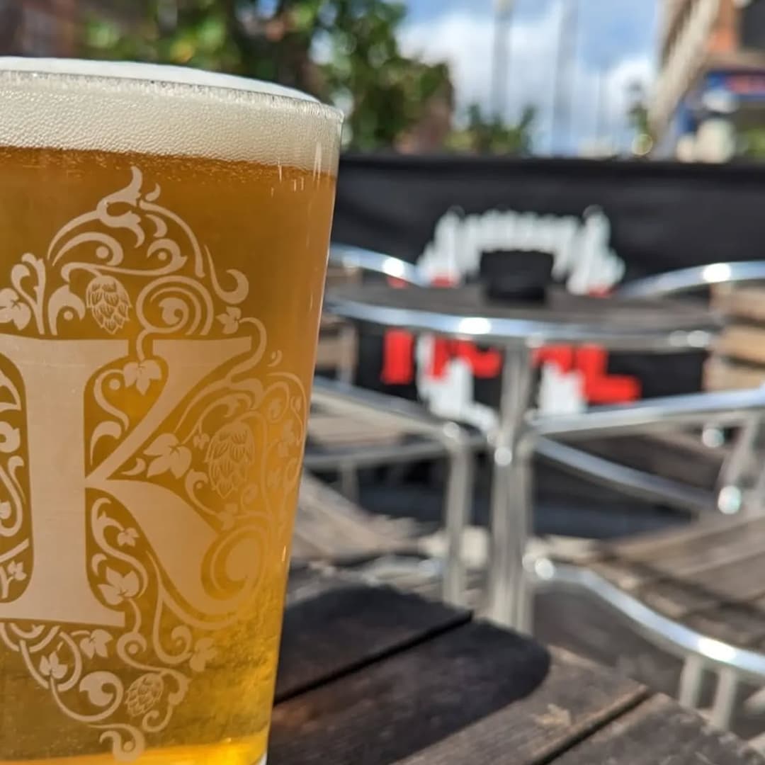 A frosty pint of beer sits on a wooden table, with a vibrant outdoor seating area in the background.