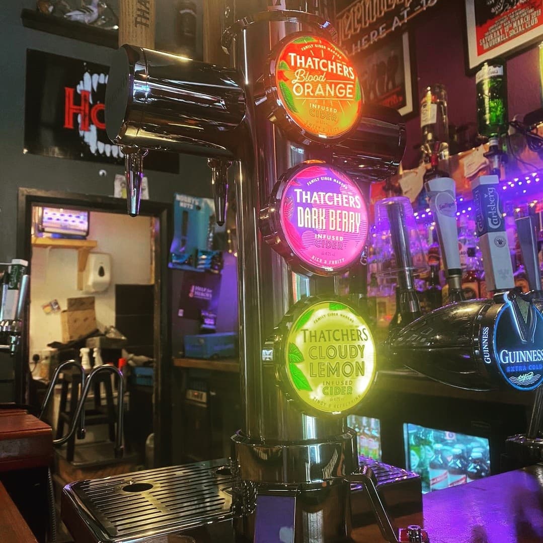 Colorful cider taps illuminated by purple lights, set against a lively bar backdrop with various drinks displayed.