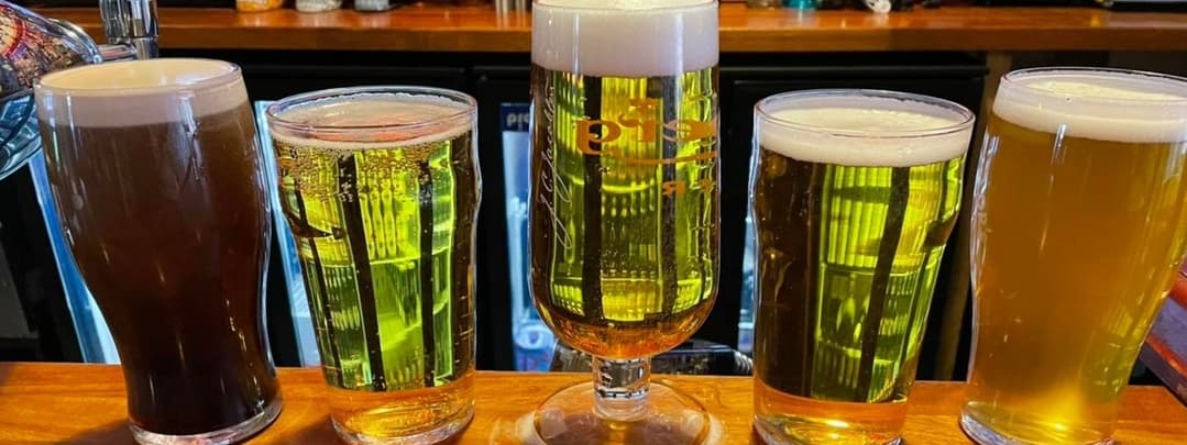 Five glasses of beer in various colors line a wooden bar, showcasing a lively atmosphere at Howl Bar Leeds.