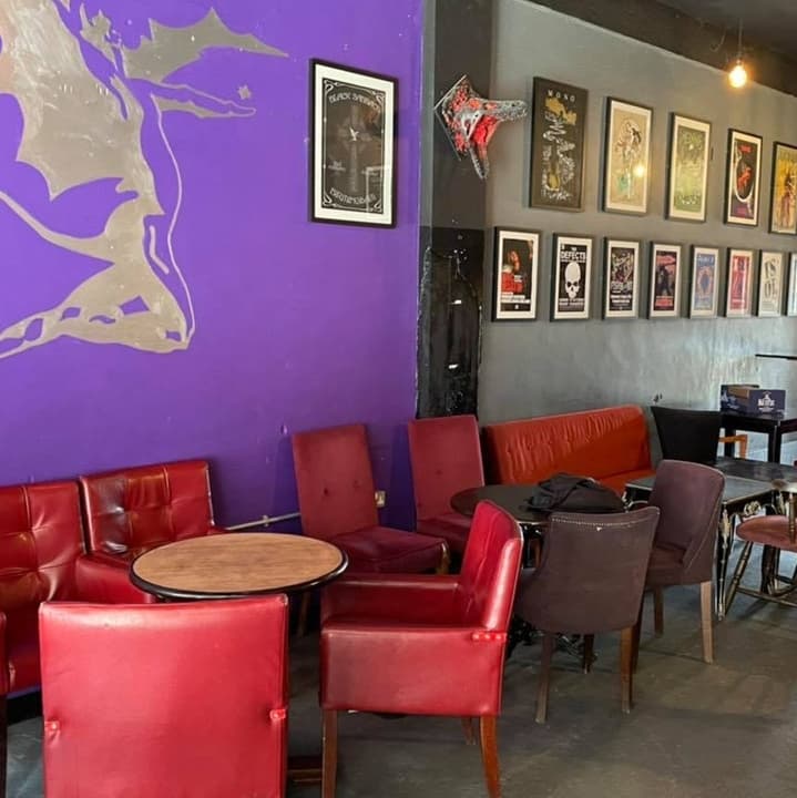 Vibrant purple wall with framed artwork, cozy red and black seating, and small round tables create a lively bar atmosphere.