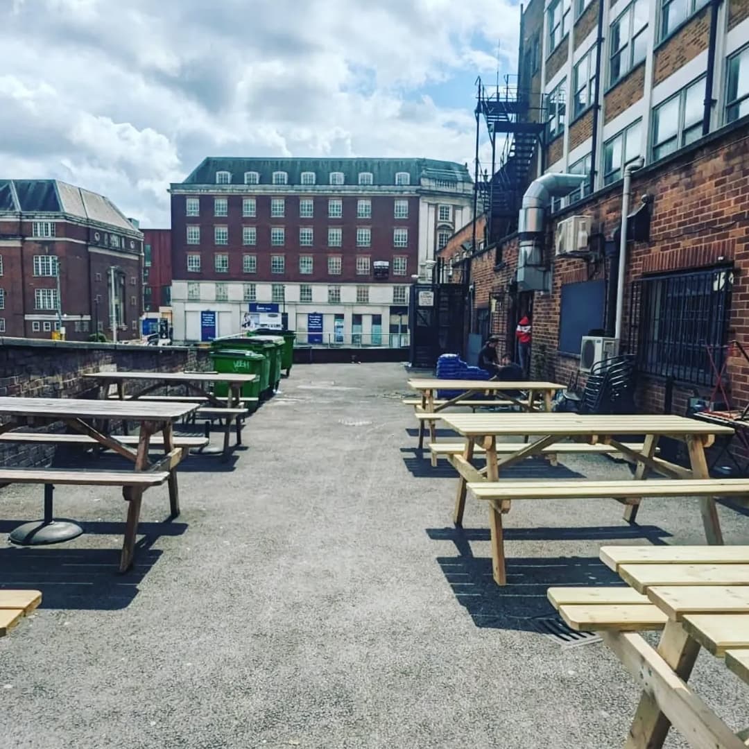 Outdoor seating area with wooden picnic tables, surrounded by brick buildings and green bins under a partly cloudy sky.