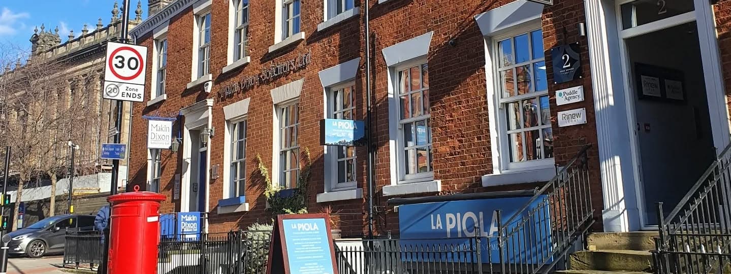 Charming brick building with large windows, outdoor seating, and a red post box, inviting diners to La Piola Italian.