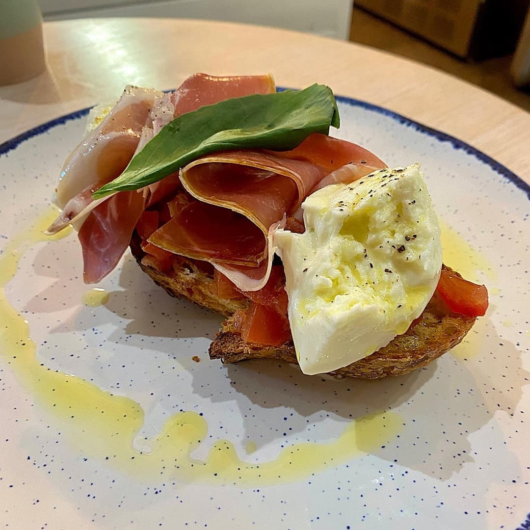 A rustic plate holds toasted bread topped with fresh tomatoes, creamy burrata, and slices of prosciutto, garnished with basil.