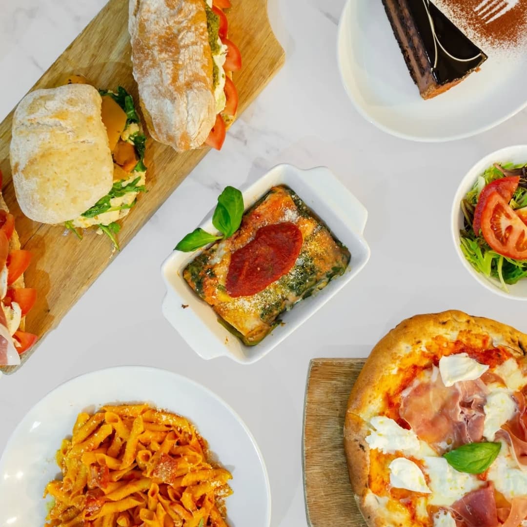 A spread of Italian dishes including lasagna, pasta, pizza, fresh salad, and sandwiches on wooden and white surfaces.