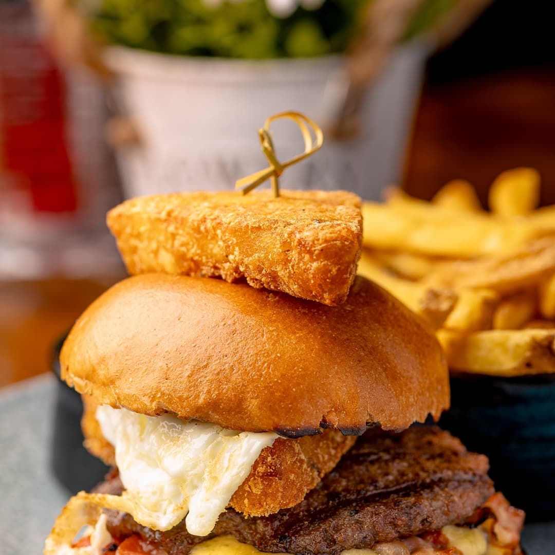 Juicy burger topped with a fried egg and crispy hash brown, served with golden fries on a rustic plate.