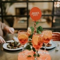 Colorful Aperol Spritz cocktails on a table, surrounded by plates of food, creating a vibrant dining atmosphere.
