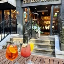 Colorful cocktails on a wooden table, with a stylish entrance to Lost & Found Leeds in the background.