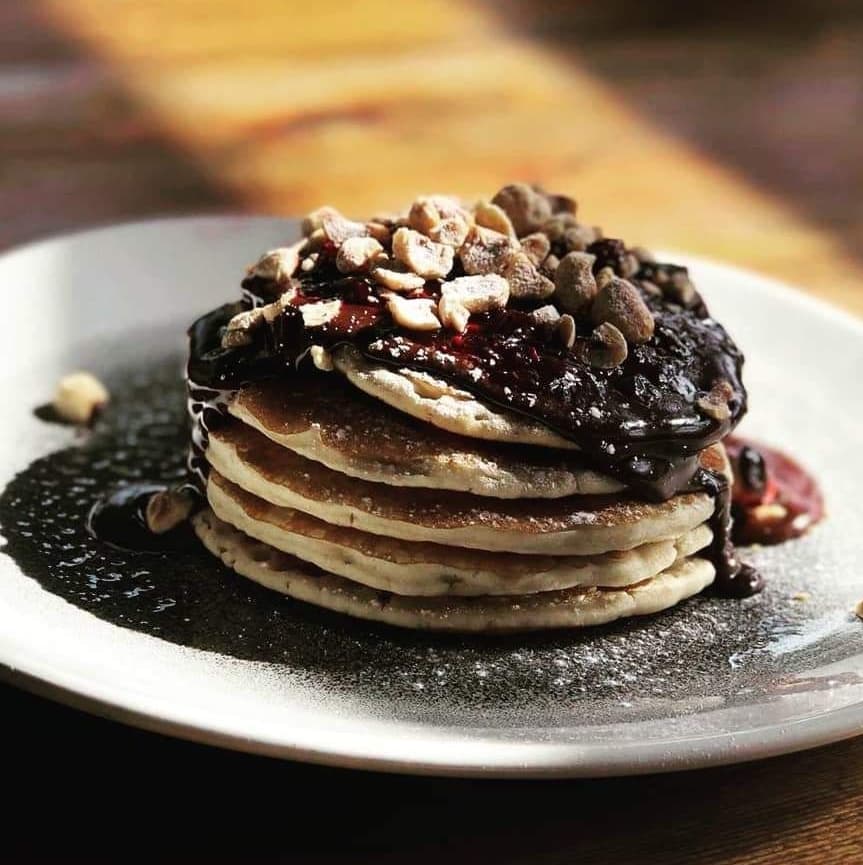 Stack of fluffy pancakes topped with chocolate sauce, crushed nuts, and a drizzle of berry compote on a rustic plate.