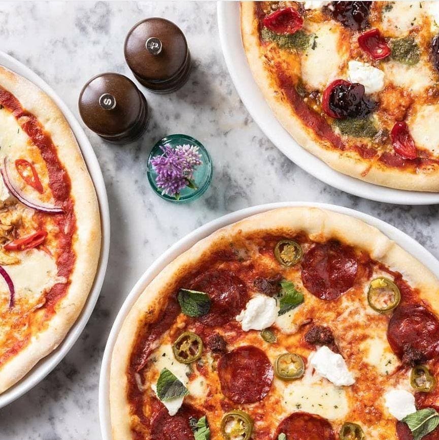 Three delicious pizzas on a marble table, topped with pepperoni, herbs, and colorful ingredients, with salt and pepper shakers nearby.