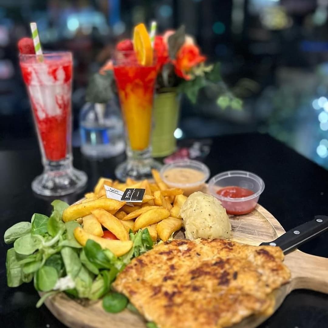 A wooden platter with grilled chicken, fries, salad, and sauces, alongside colorful cocktails in tall glasses.