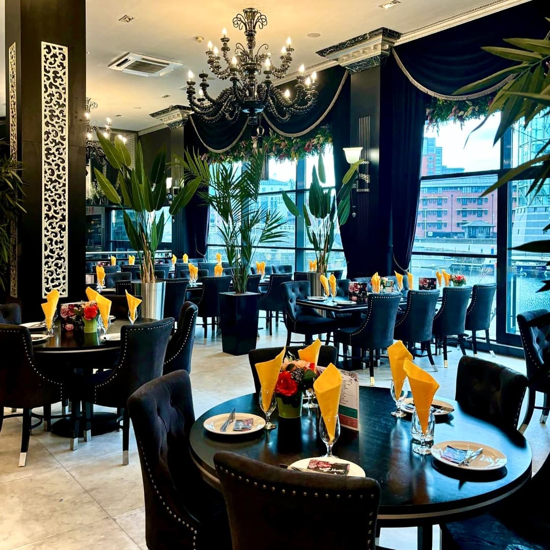 Elegant dining area with black chairs, yellow napkins, and floral centerpieces, featuring large windows and chandeliers.
