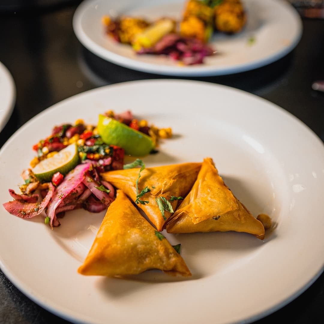 Three golden-brown samosas on a white plate, accompanied by a colorful salad with lime and herbs.